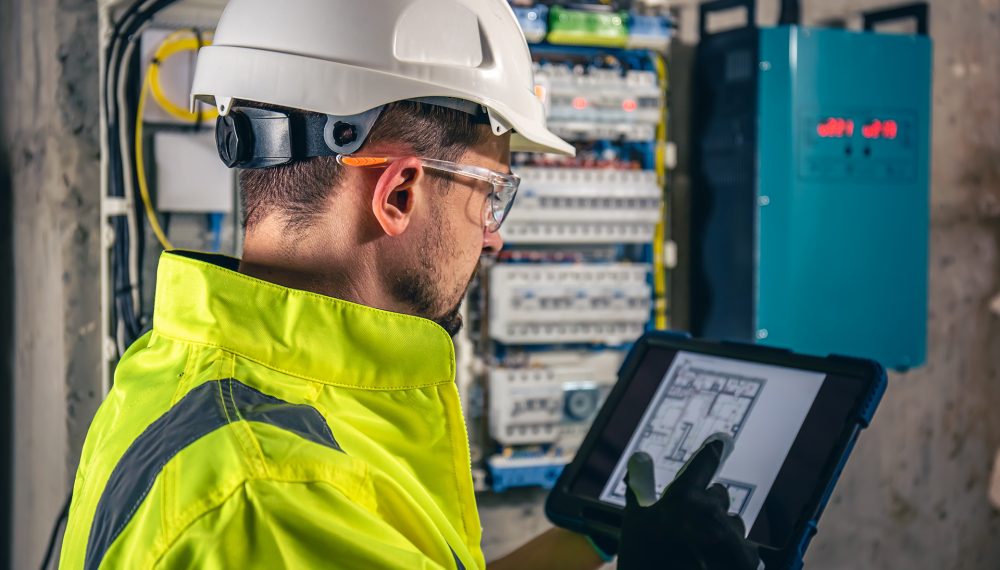 Man, an electrical technician working in a switchboard with fuses, uses a tablet. Man, an electrical technician working in a switchboard with fuses. Installation and connection of electrical equipment. Professional uses a tablet.